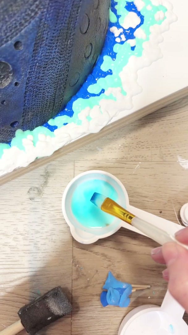 Studio process clip: turquoise and blue paint piped into a reef-like ridge around the dark encaustic dome of Eye Sea All I, a brush loading colour from a cup