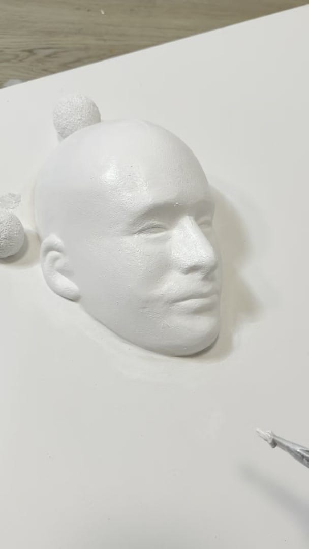 Studio process clip: a white sculpted relief of a human face on a white panel, small white spheres beside it