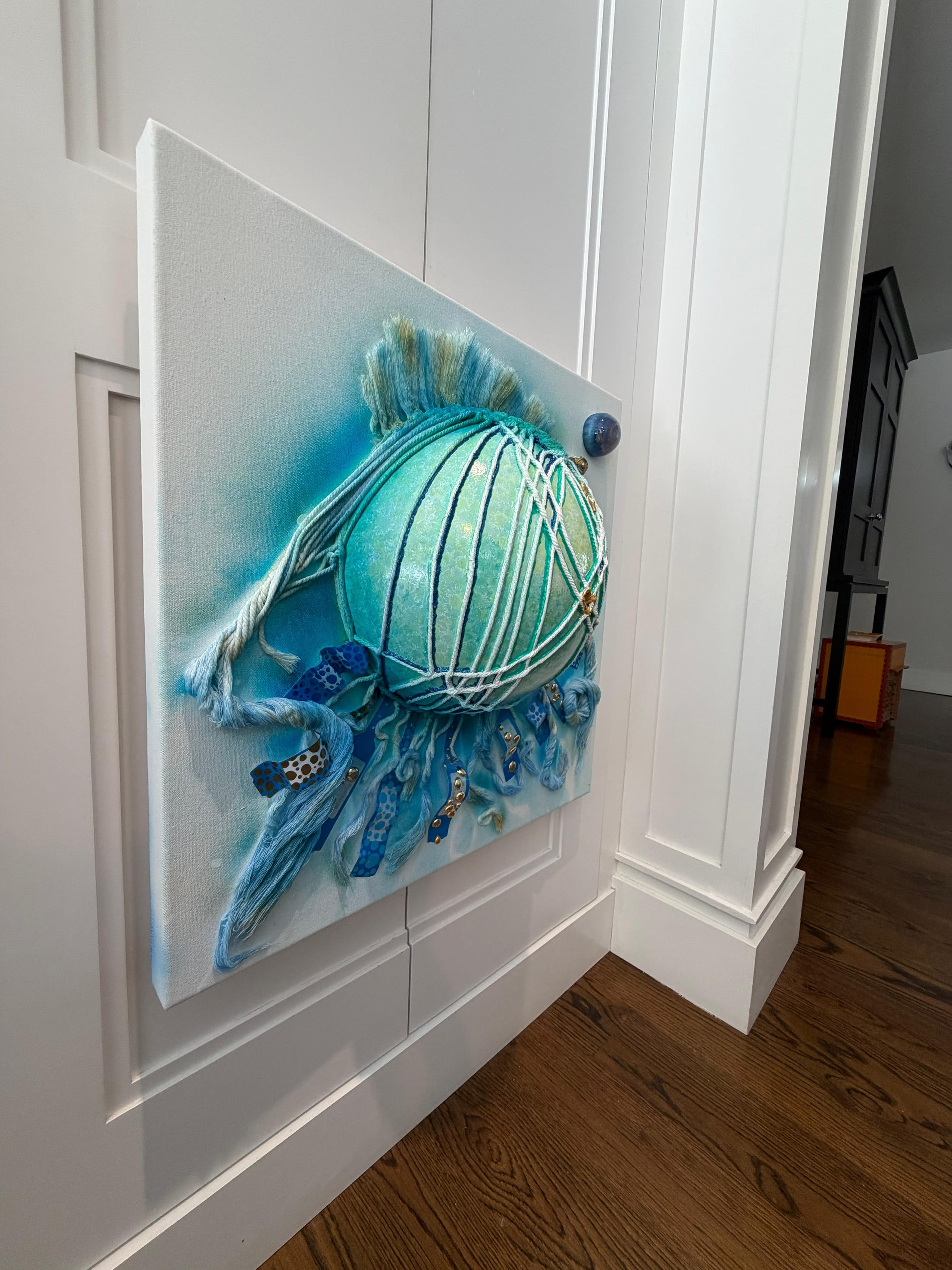 Faceless Self Portrait by Victoria Zeder installed on a white panelled wall: a netted teal encaustic sphere with a fibre crest and hanging immobilization-band tentacles, seen at an angle