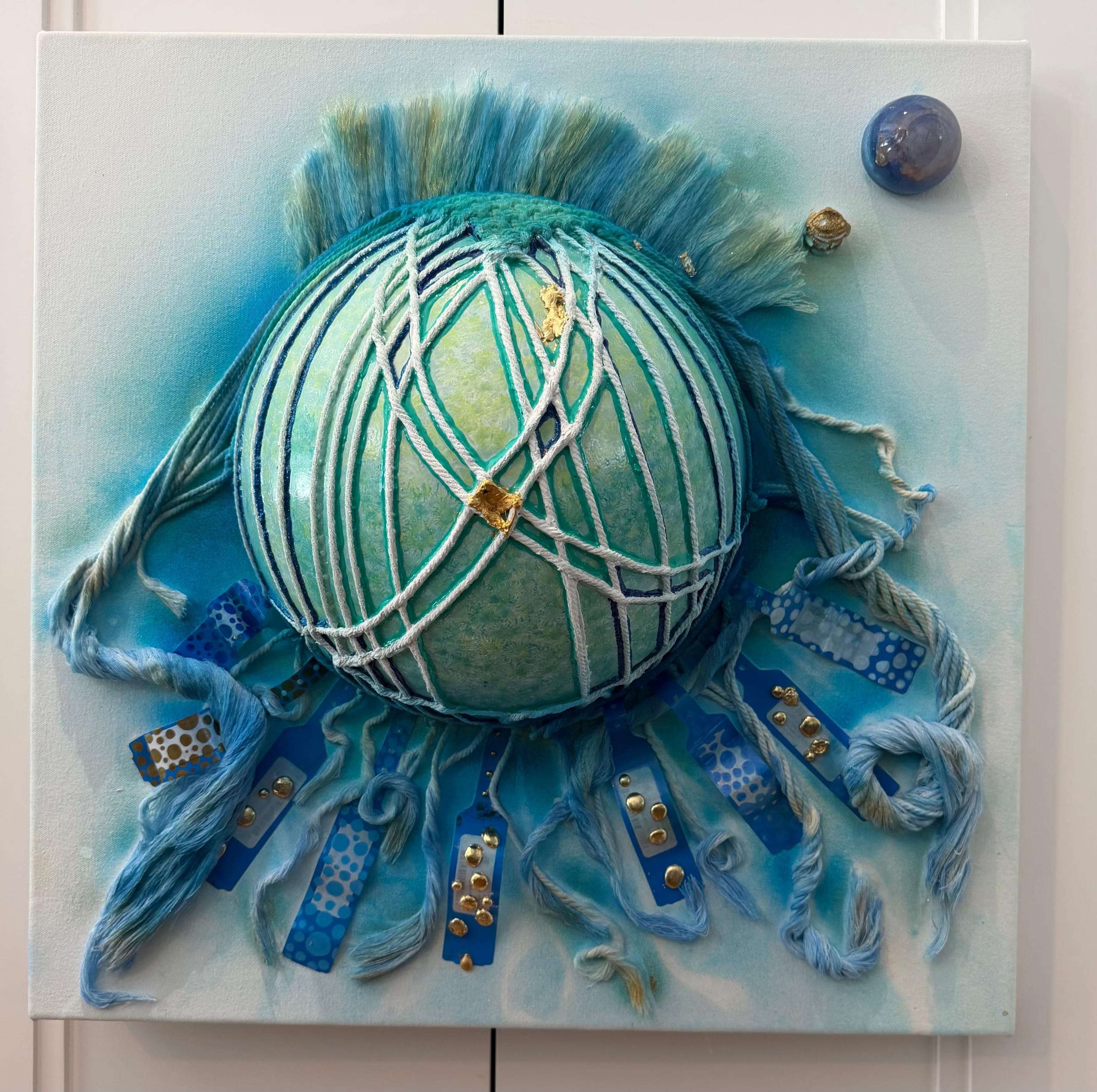 Faceless Self Portrait by Victoria Zeder seen straight on: a netted teal encaustic sphere with a fibre crest and blue immobilization-band tentacles studded with gold, on cerulean canvas