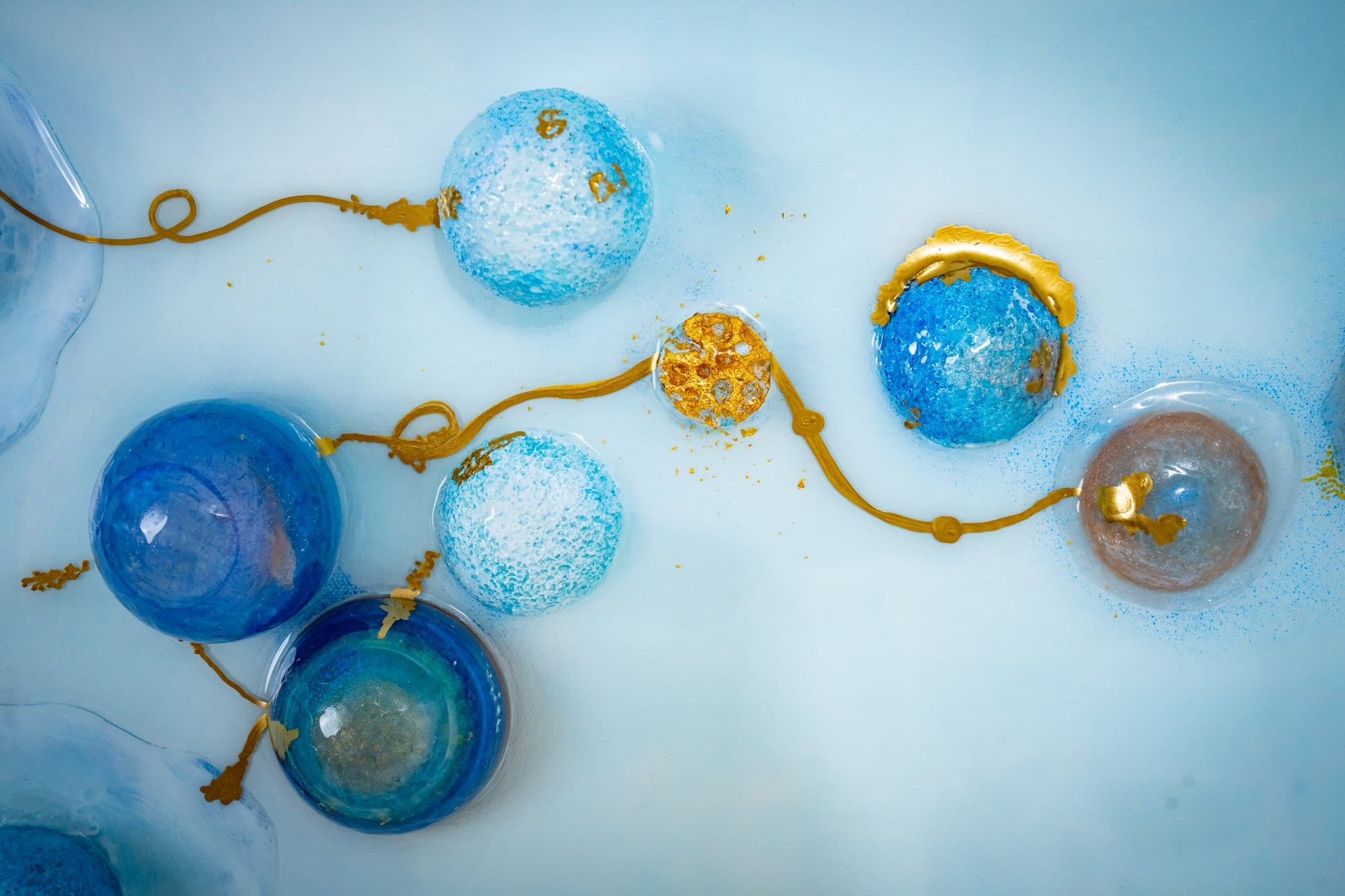 Macro from the Origin sub-series: translucent blue resin spheres of varying tone linked by a meandering gold filament across a pale cerulean field