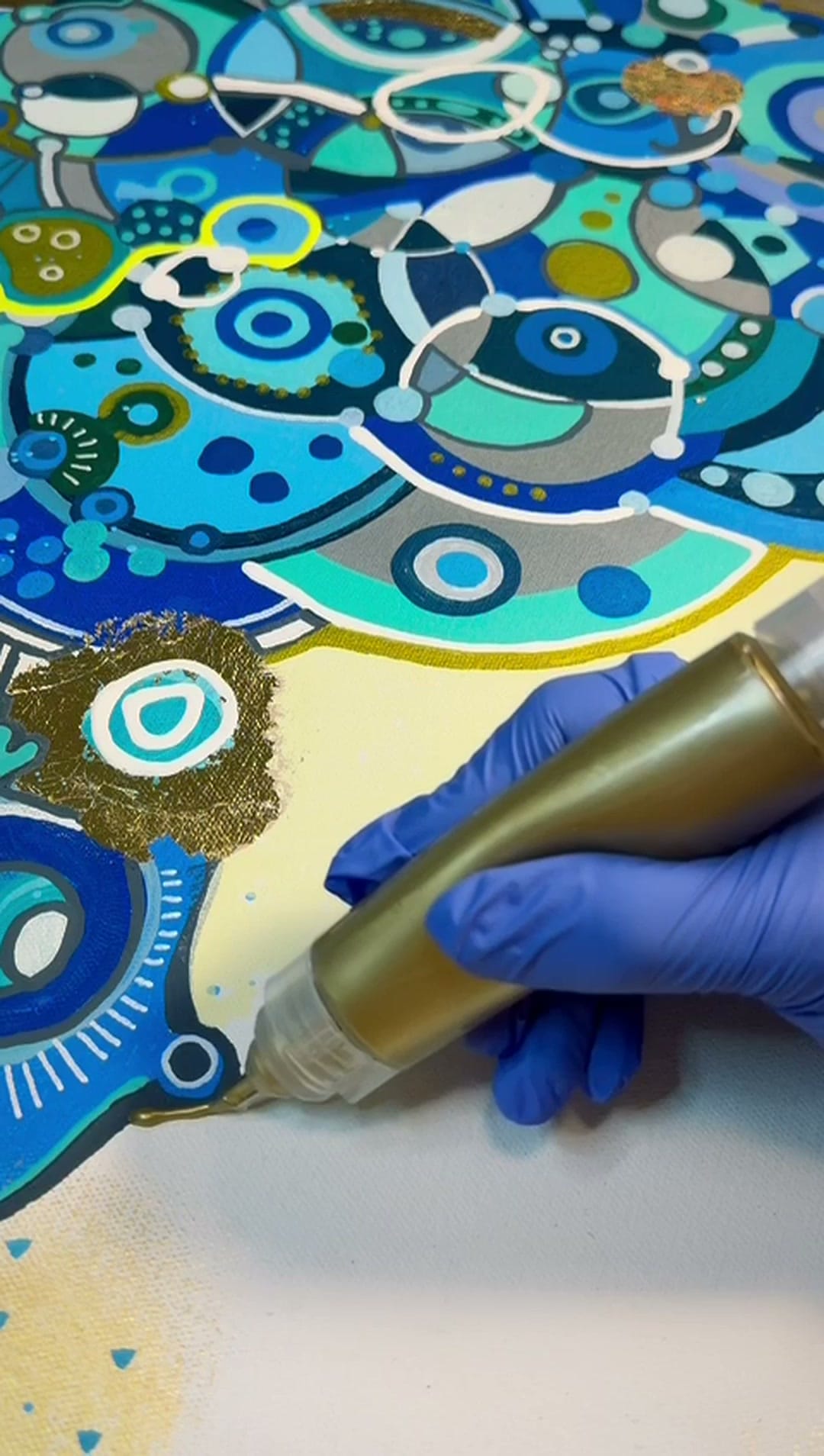 Gloved hand holding a gold paint pen, approaching a cobalt and grey painted cluster on cream canvas