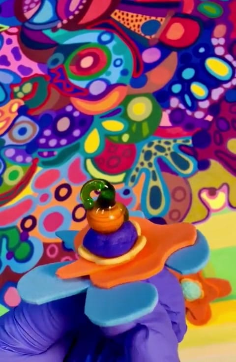 Studio process clip: a gloved hand holding a small colourful sculptural bead up against a vivid psychedelic multicoloured painting